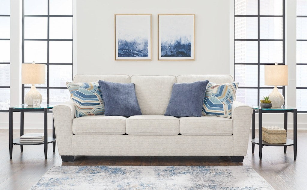 Full front view of the sofa in a light, textured fabric (light beige/oatmeal), beautifully staged in a modern living room setting with two matching end tables, lamps, and abstract blue artwork.
