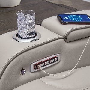 Close-up shot of the sofa's armrest, revealing an illuminated cup holder, multiple USB charging ports (Type-A and Type-C), a power outlet, and recline controls.